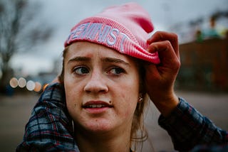 Young girl having a pink beanie placed on her head reading “FEMINIST” across the front.