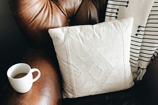 A cozy image of a brown leather armchair holding a cup of coffee and a white pillow