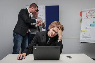 A person with a pained expression and one hand on their head sits in front of a laptop. People in the background discuss some documents.