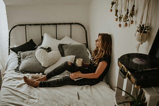 A person sitting in their bed with a cup of coffee or tea. They look tired.