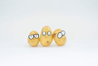 Three small potatoes lined up in front of the camera. They are in the center of the image and each one is wearing a paper cut out with fake eyes stickered onto the top half of the potato. The first one is wearing glasses and their eyes are shut the second looks surprised and the third looks confused