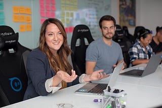 image shows a career woman in a blazer explaining something to someone across a white boardroom table. In front of her is a laptop. Behind her are post it notes on a glass wall.