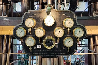 An elaborate old instrument panel with 9 circular dials and a counter, all with brass surrounds on a black cast iron plaque. Behind a complex network of pipes and machinery
