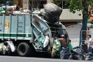 Garbage truck driving down the street, loaded with trash bags, symbolizing the burden of unchecked anger spilling over and leaving a trail of mess in its path