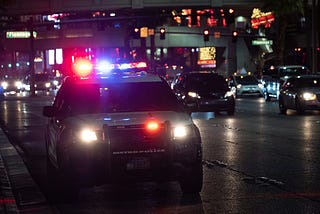A police car with flashing lights is parked on a busy city street at night, surrounded by traffic and illuminated signs.
