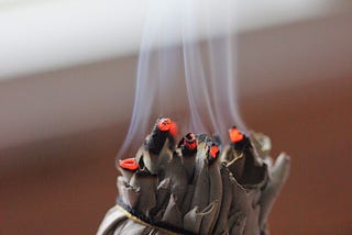 A close up photograph of a sage bundle, lit at one end with smoke rising from the ignited leaves.