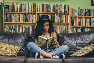 Woman with long dark hair and a black hat on sits cross-legged on a couch reading. Full bookshelves line the wall behind her.