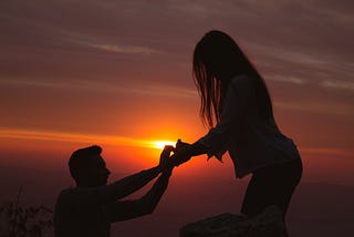 A man proposes to his girlfriend, kneeling before a beautiful sunset.