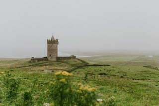 A small stone tower sits on a hill on a coast. The sky is foggy.