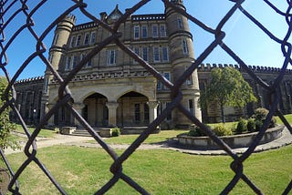 Looking through the metal fencing towars the large, stone building with turrets. Theres a grassy lawn and some trees in front of the building.