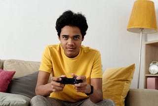 A man in a yellow shirt holds a black video game controller. He is focused intently on the game and will surely level up his progress bar soon. He sits on a couch. The couch has a red and grey chushion. There is also a yellow cushion and a yellow lamp shade on a tall standing lamp behind the man. They compliment his yellow shirt well. To the right of the frame is small bookcase with a gold alarm clock and a pink block. The man has black hair that is somehwat unkempt, as such a gamer would have.