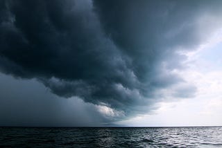 A giant cloud over the ocean, on the right side you can see the sunlight peeking through the clouds, on the left side the sky is dark.