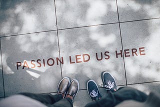 Shoes and legs of two people are visible with text printed on floor “Passion Led Us Here”