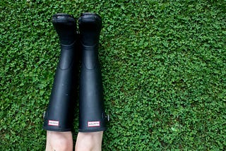 On a field of emerald green clover a pair of black Hunter Wellington Boots lie souls to the top edge of the picture. Two very white knees stick out of the boots at the very edge of the bottom of the picture.