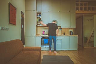 A man in blue jeans and black t-shirt stands, back facing the camera, over his kitchen counter, waiting for the coffee to boil