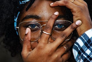 An african american woman with curly hair, a hand covering her face, with vibrant blue eyes, and a blue and white plaid shirt and bandana to match.