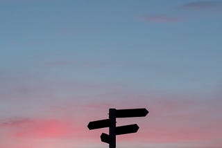 A signpost stands silhouetted against the sky at sunset.