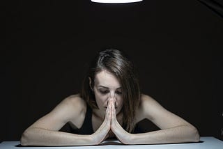 A woman seated, elbows on the table, hands joined at face level, eyes closed, worried atmosphere