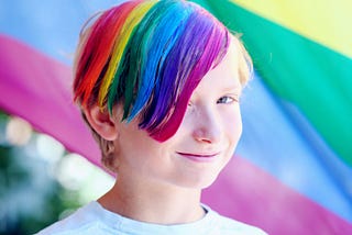 A smiling child with hair dyed in a rainbow pattern, in front of a rainbow flag.