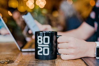 A person holds a mug placed on a desk while looking at their phone with their laptop in front of them.