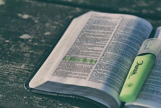 An open book filled with text, with a yellow highlighted passage on the left page. In the center of the book lies a yellow highlighter pen.