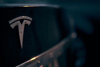 A close up picture of the Tesla logo on the back of a Tesla automotive vehicle.