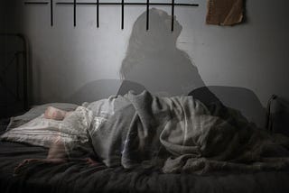 A woman lays in a bed barefoot, covered by a sheet. A second image is less saturated layering the first: a woman sits up in bed looking up. She has long hair and is wearing a dark sweater.