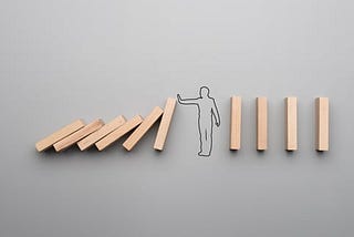 A number of domino-like pieces of wood. The left half has fallen over, and the right half is standing. A line drawing of a person is in between these two halves, preventing the falling piece of wood from hitting the next standing one.
