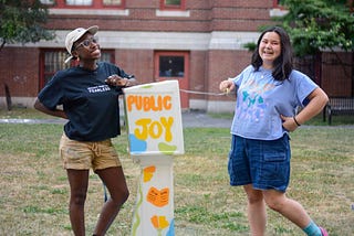 Sabrina and Ava stand with a four-foot-tall box between them on a patch of grass. They are both smiling, and Ava opens the top of the box. The box is painted with bright orange, yellow, green, and blue colors and says the words “Public Joy”.