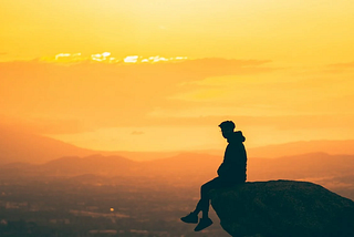 Silhouette of a boy sitting on a rock overlooking a valley that is drenched in the orange glow of sunset.