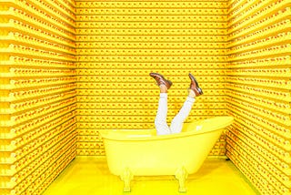 The LUnatic Poet headfirst into a yellow tub, feet sticking up in the air. He is surrounded by shelves full of rubber ducks.