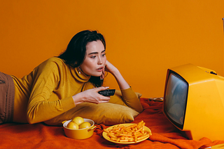 A woman in a yellow long-sleeved shirt lying on her side while watching something on a Yellow TV
