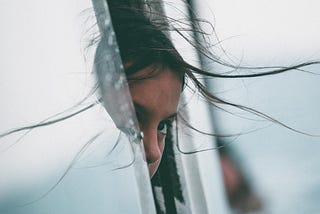 A young girl hiding behind a glass door with windswept hair.