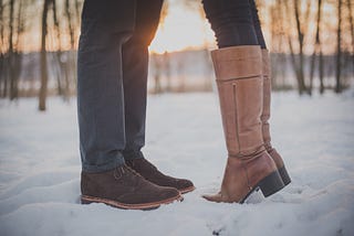 The legs and feet of a man and woman who appear to be kissing. The woman is standing on her tip toes in knee-high boots.