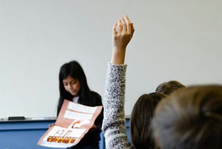 A student raises her hand as her classmate makes a poster presentation in the front of the room.