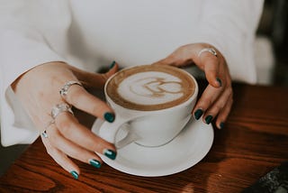 hands about to grasp a white mug of coffee with latte art on top of it