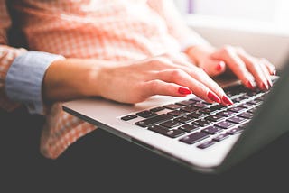 A white woman with red fingernail polish types on a Mac laptop, which sits on her lap.
