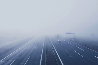 A multi-lane motorway disappearing into dense fog, with only a few vehicles visible and road markings fading into low visibility.