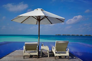 Two beach loungers on a desk, under a big white umbrella overlooking pristine blue waters.