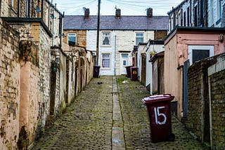 A narrow alley between old terraced houses. Paint crumbles from the bricks, and the neighborhood looks run down.