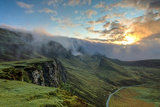 wide open vista, with craggy hills covered with green, a river in the distance, and sun peeking out behind clouds