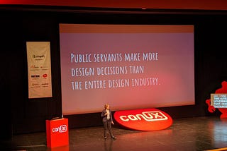 A woman on a stage standing in front of a slide that says ‘public servants make more design decisions than the entire design industry’