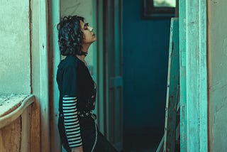 A woman in a black shirt and jeans with a striped undershirt leans against the doorframe of a rundown apartment.