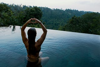 A woman in a bathing suit sitting in an infinity pool that is overlooking a lush green jungle. She is also making a heart sign with her two hands.