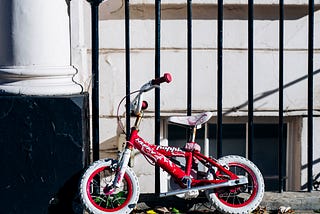 A pink child’s bike without training wheels propped against a wrought iron fence in a city