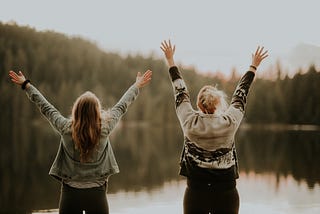 Two women facing away from the camera. They have their hands up in celebration while looking at a river.