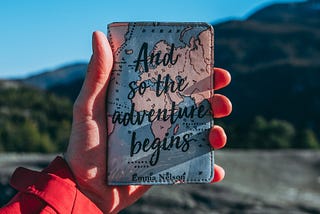 A hand is holding up a passport holder with the phrase “And so the adventure begins.” In the background is a blurred mountain terrain with a clear blue sky.