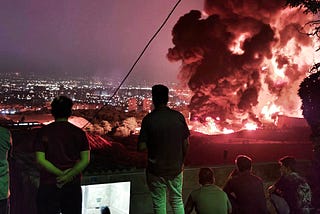 People silhoutted against the night sky, watching a huge fire against the dim lights of the city