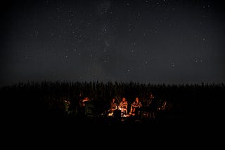 People gathered around a fire at night. Behind them the black silhouette of a treeline. Above them the starry night sky.