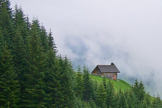 A hut perched on the side of a mountain in a small clearing and surrounded by fir trees, overlooking clouds and mist.